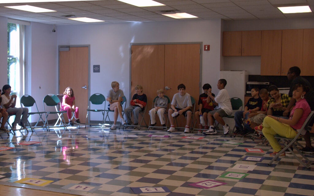 A group of children and an adult sit in a circle of chairs in a classroom with number cards arranged on the floor.