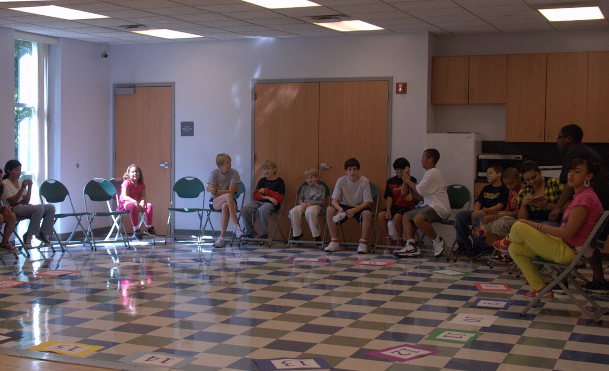 A group of children and an adult sit in a circle of chairs in a classroom with number cards arranged on the floor.