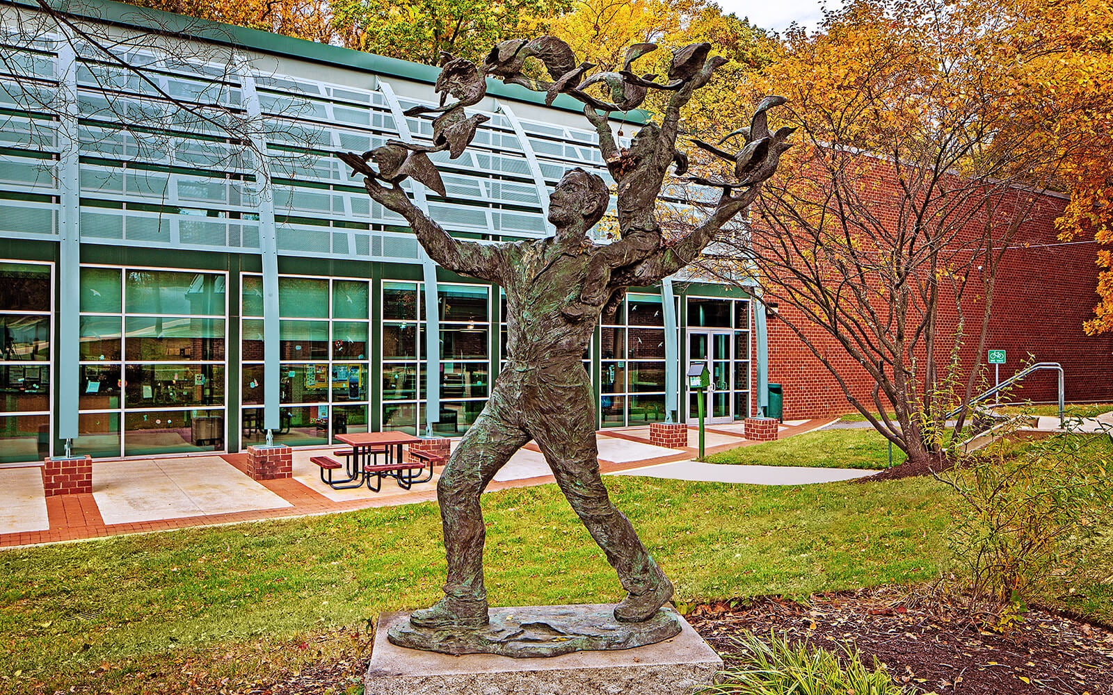A bronze statue of a person releasing multiple birds stands on a stone base in front of a modern glass building with a brick exterior and outdoor seating.