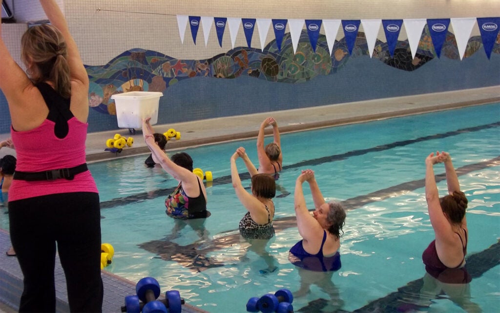 A group of adults in a swimming pool perform stretching exercises led by an instructor at the poolside.