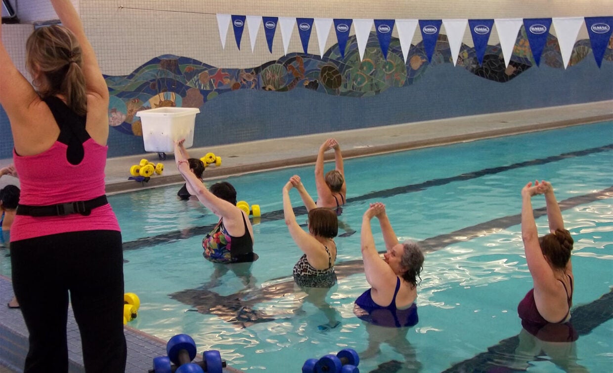 A group of adults in a swimming pool perform stretching exercises led by an instructor at the poolside.