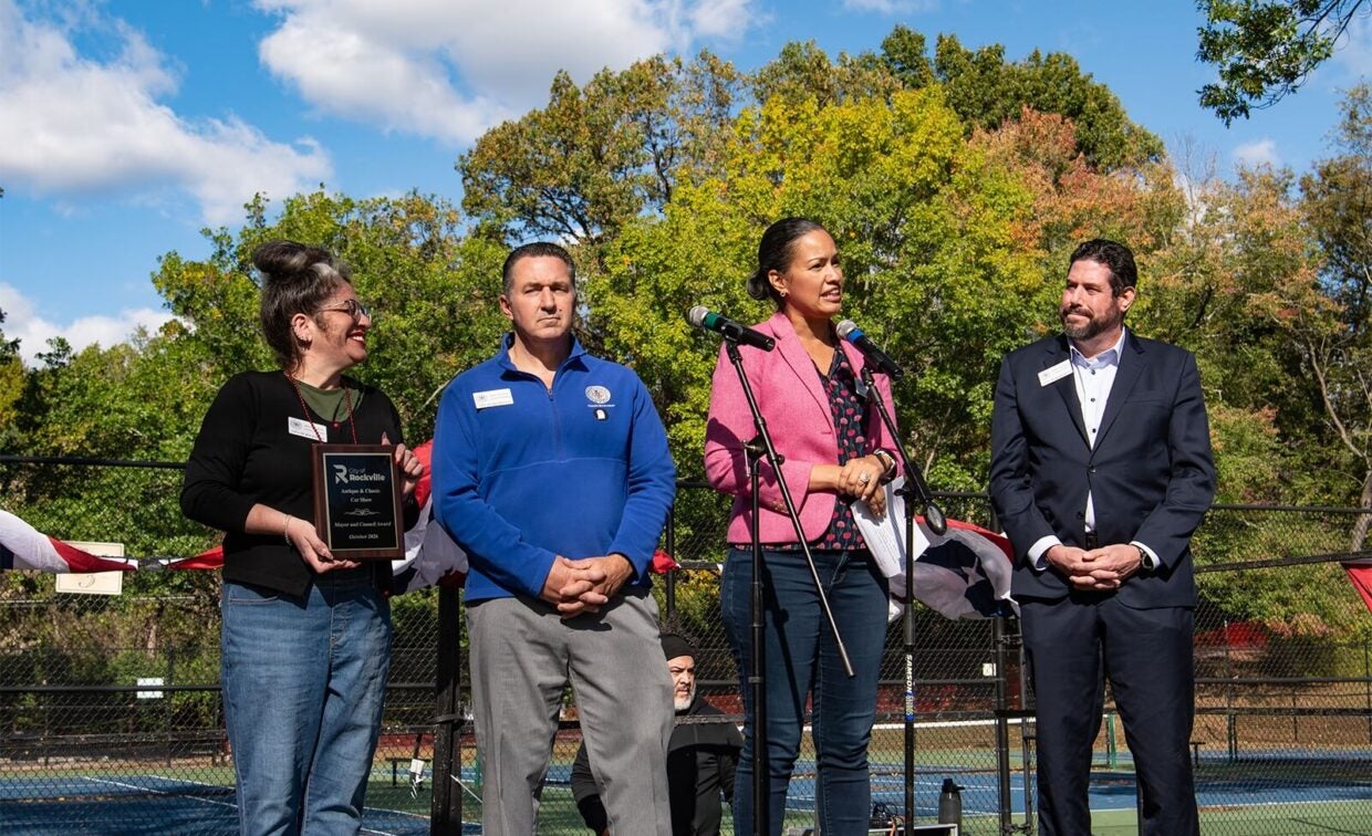 Four people stand on a stage outdoors; one woman holds an award plaque while another speaks into microphones.
