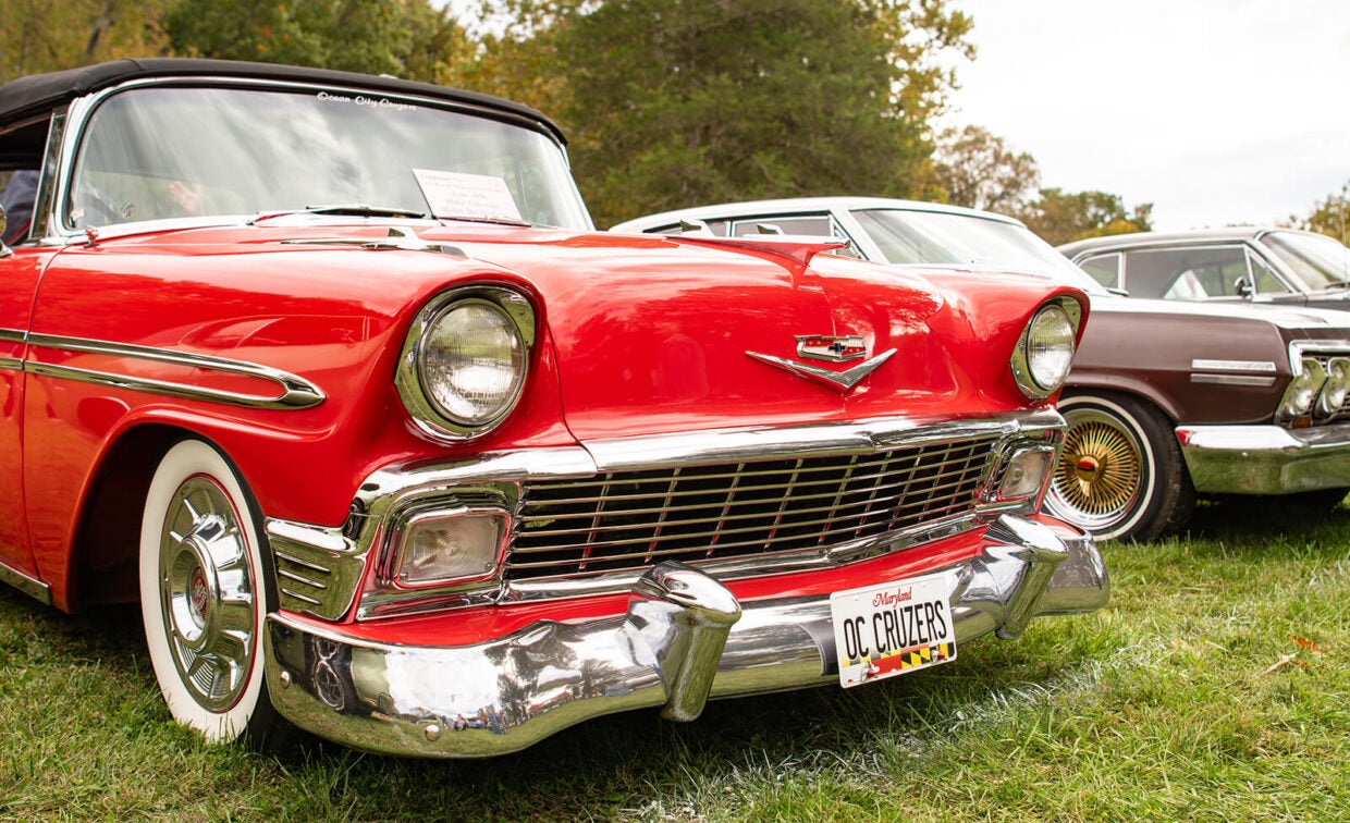A classic red Chevrolet with a chrome bumper and a Maryland “OC CRUZERS” license plate parked on grass next to other vintage cars.