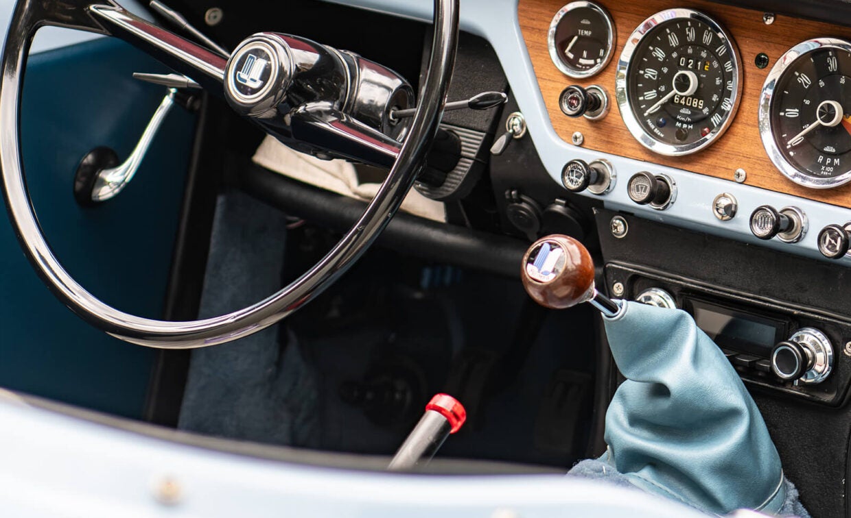 The driver’s side interior of a vintage car, showing a steering wheel, a wooden gear shift knob, and a wood-grain dashboard with round analog gauges.