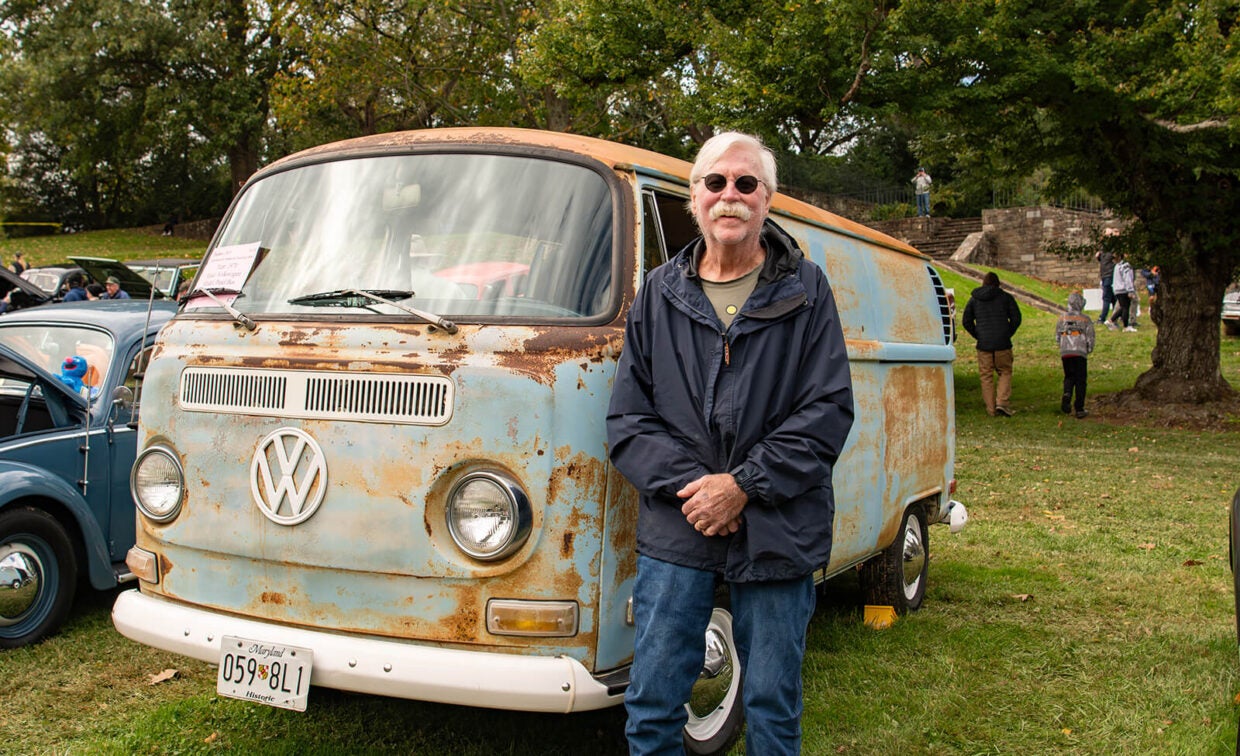 A man with a mustache stands in front of a rusted light-blue vintage Volkswagen van at an outdoor car show.