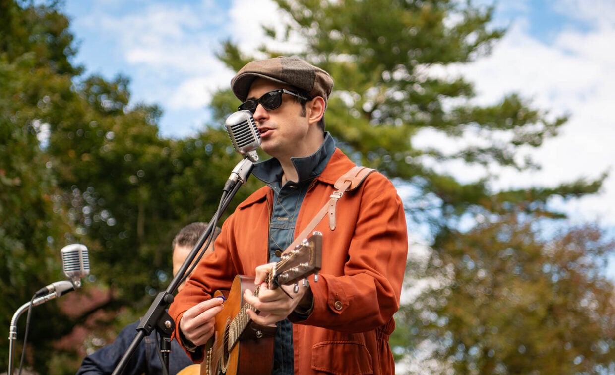 A musician in a cap, sunglasses, and an orange jacket plays an acoustic guitar while singing into a vintage-style microphone.