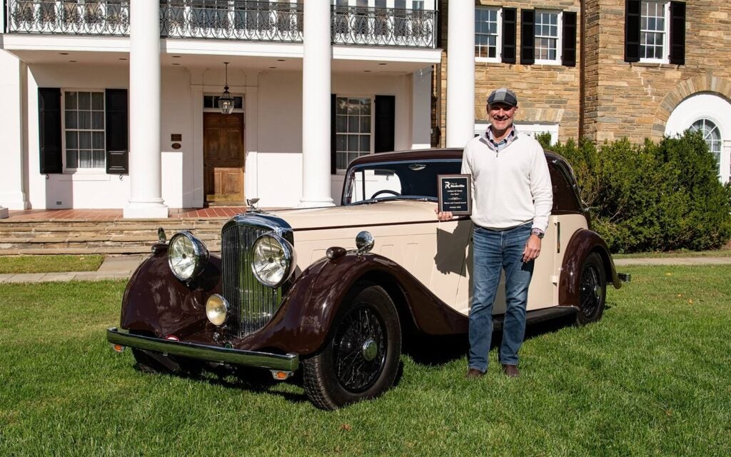 A man stands next to a two-tone vintage sedan in front of a colonial-style mansion, holding a plaque from a car show.