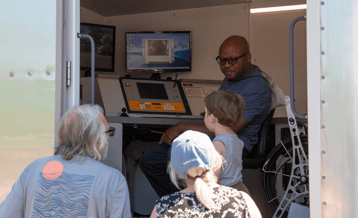 A man sits at a control console inside a mobile workstation, showing pipe inspection camera footage on a monitor to two adults and a child.
