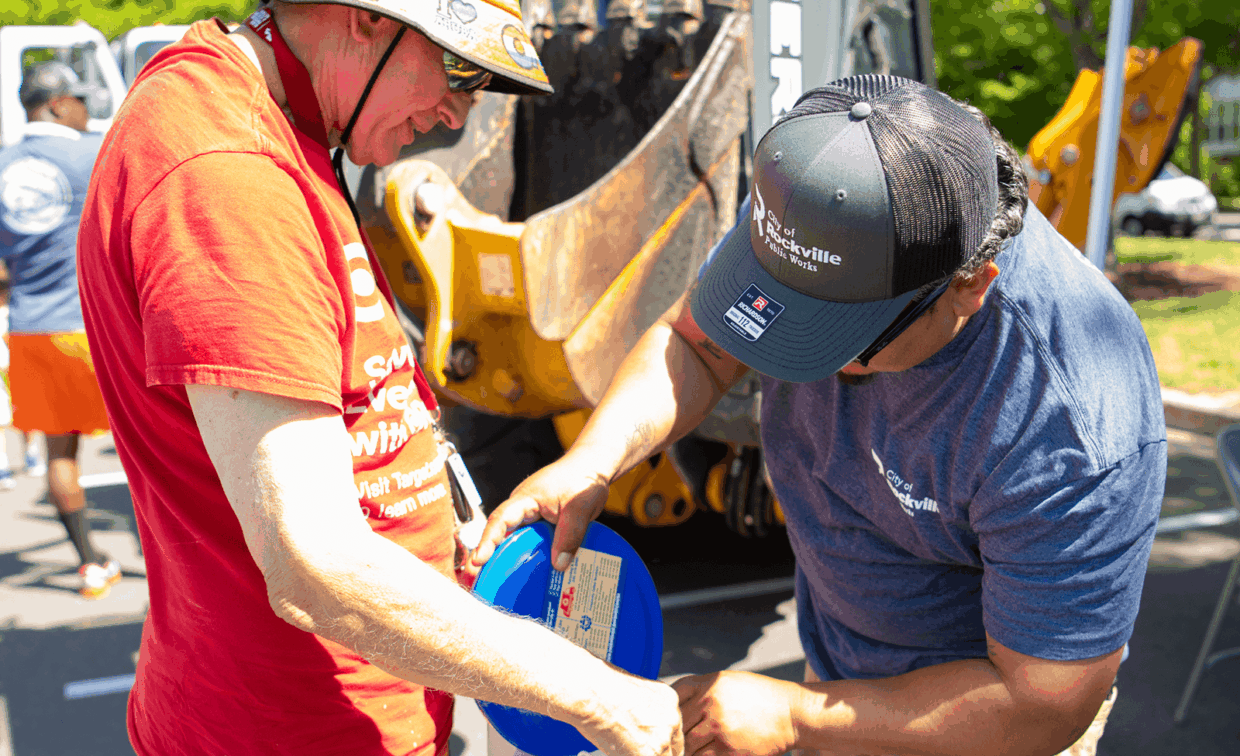 Two workers in Rockville Public Works shirts examine a blue plastic container near a piece of yellow heavy machinery.