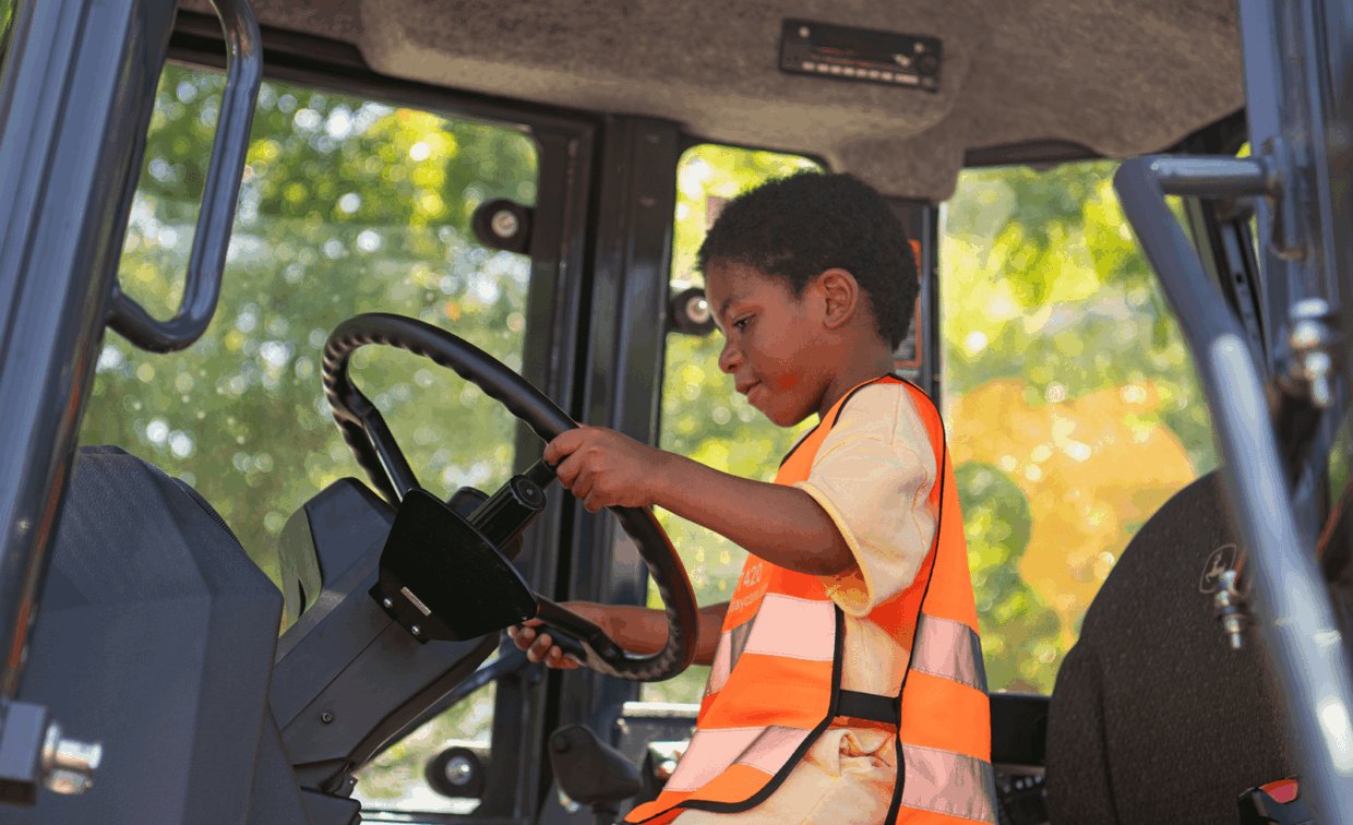 A child in an orange safety vest sits in the driver’s seat of construction equipment, holding the steering wheel.