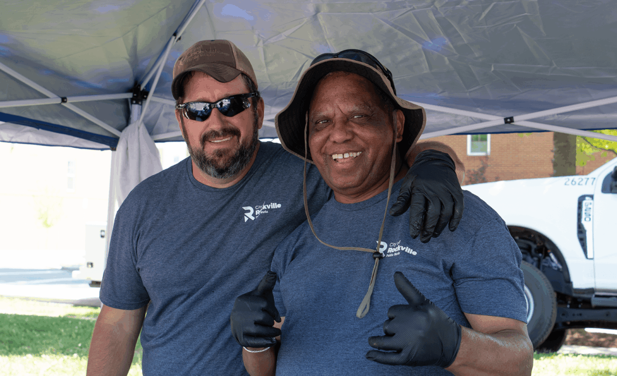Two City of Rockville Public Works employees in matching t-shirts and black gloves smile under a canopy tent.