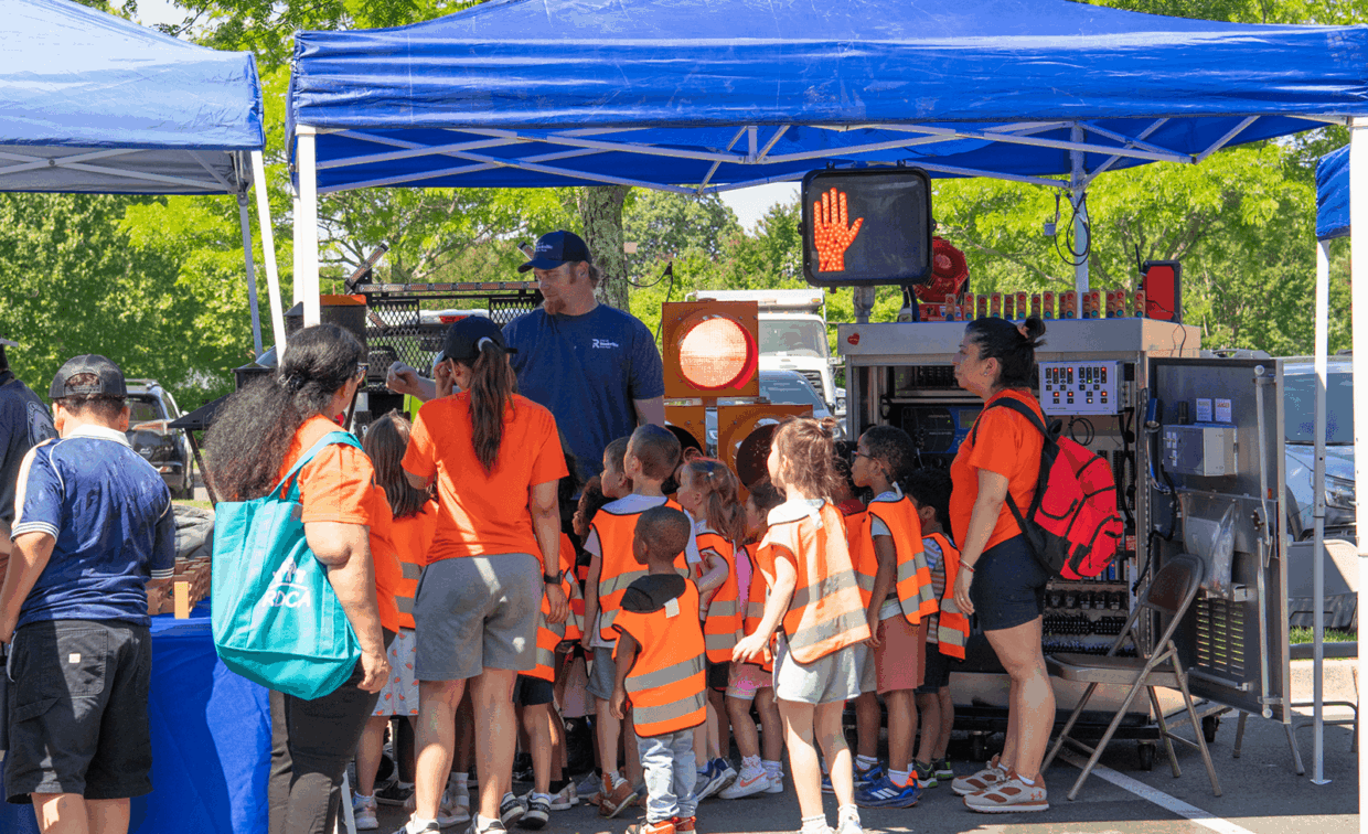 A group of children in orange safety vests look at a traffic signal display under a blue canopy.