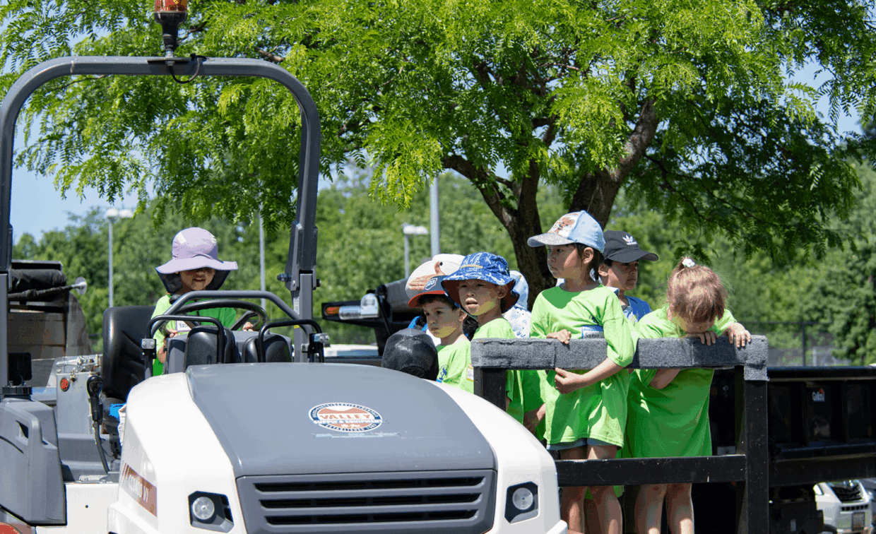 A group of children in lime green shirts standing on the back of a utility vehicle under a tree.