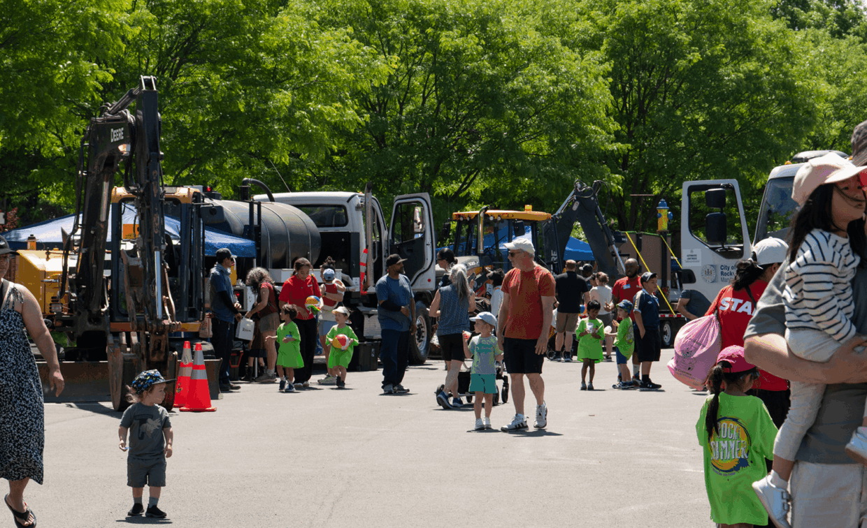 Children and adults gather around construction vehicles and work trucks at an outdoor community event.