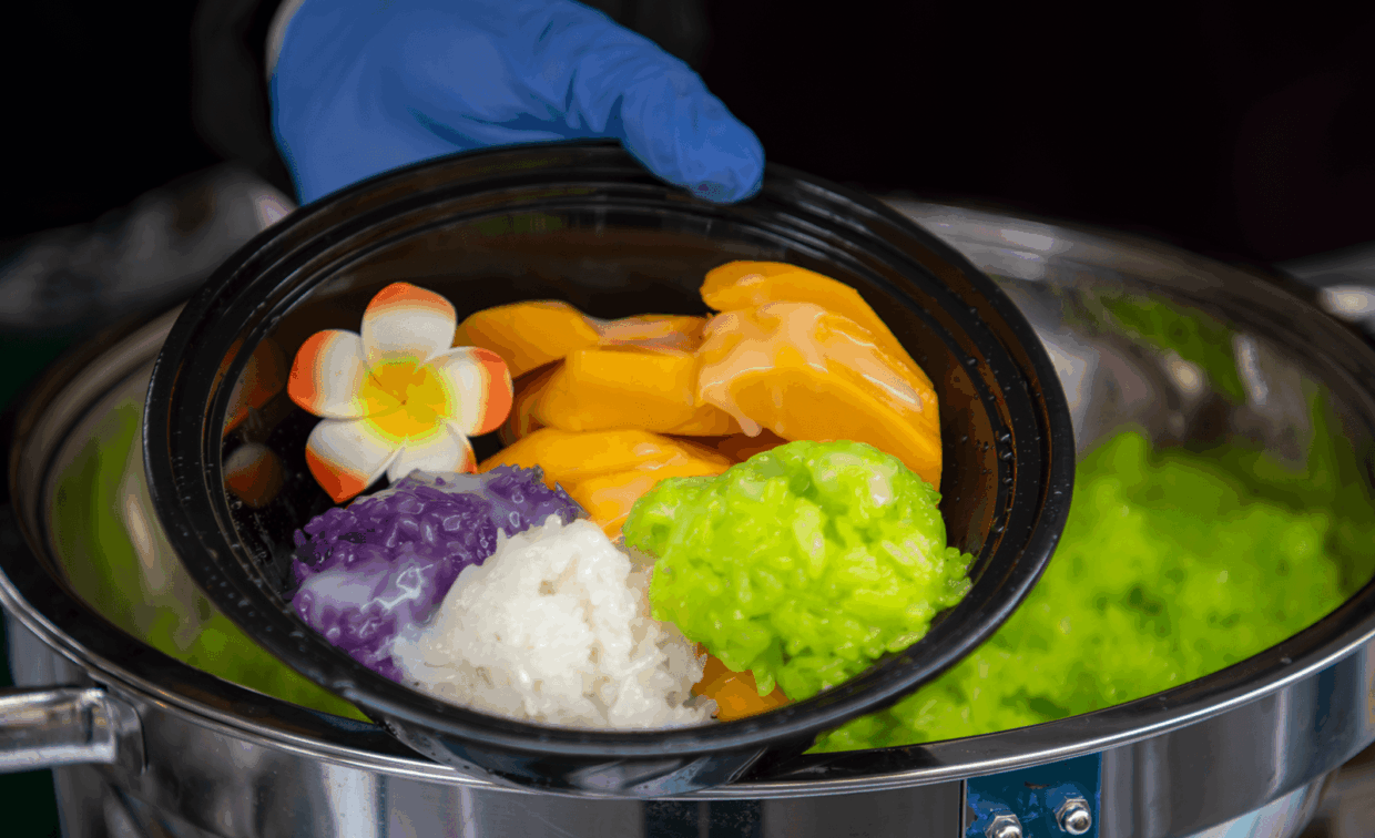 A hand in a blue glove holds a bowl of mango sticky rice featuring purple, white, and green rice, garnished with a flower.
