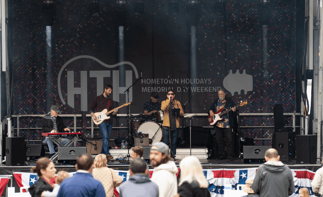 A live band performs on a stage with a Hometown Holidays banner, seen from behind an audience.