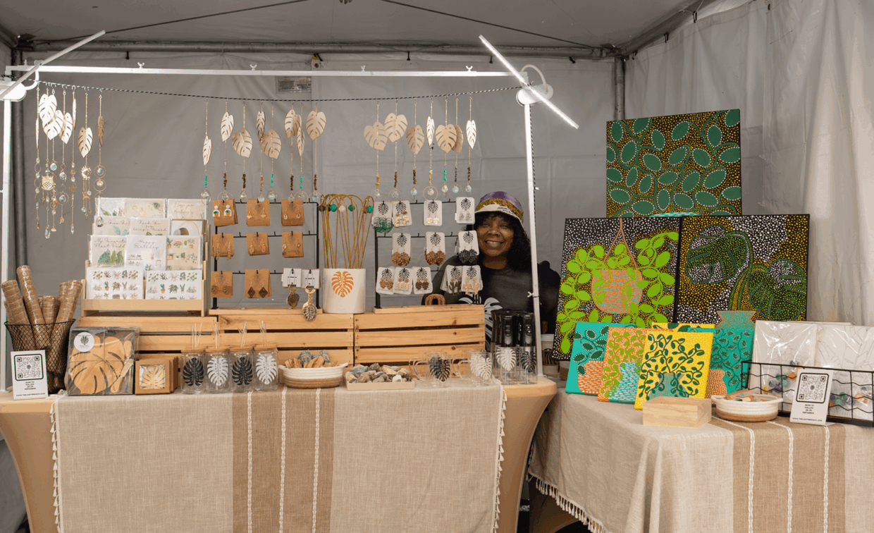 A vendor stands behind a table at an outdoor market, displaying hanging wooden earrings, greeting cards, and patterned dot-art paintings.