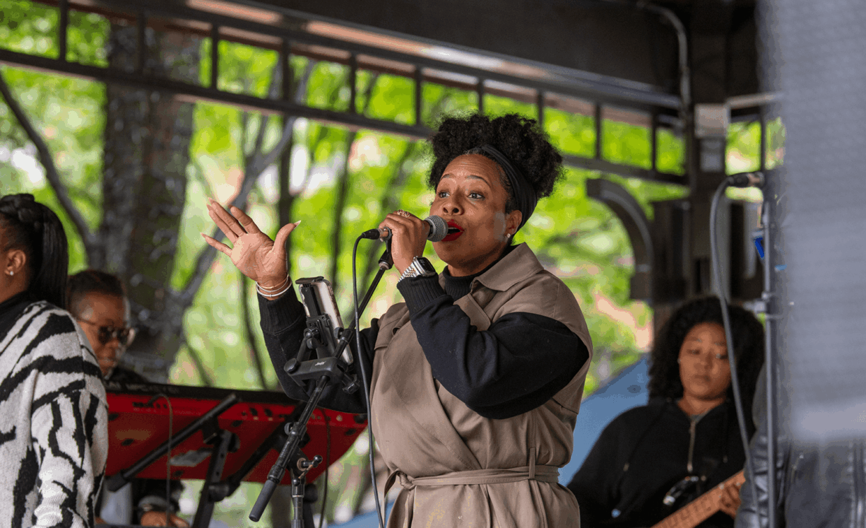 A woman singing into a microphone on an outdoor stage with band members in the background.