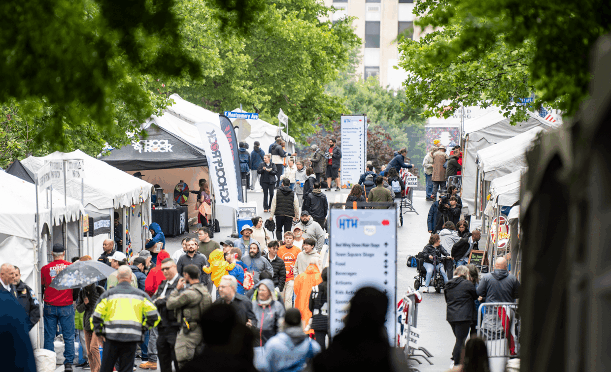 A crowded outdoor street festival with white vendor tents, event signs, and people walking along the street.