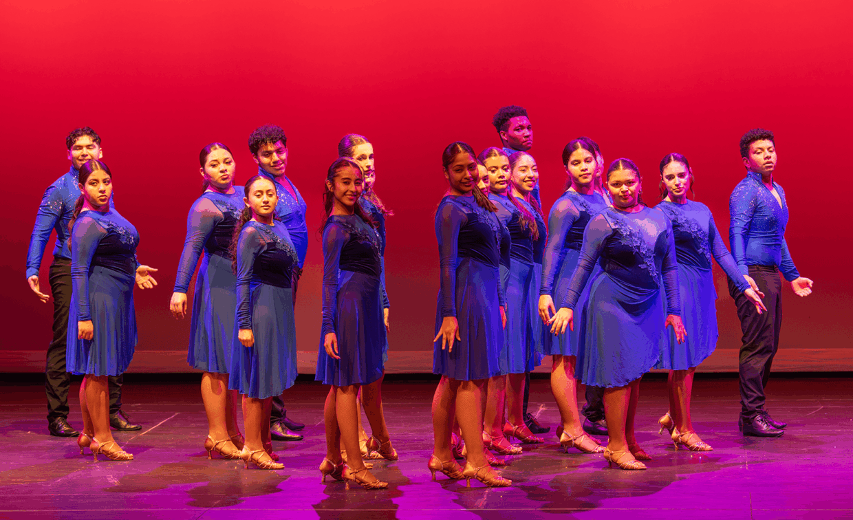 A group of dancers in matching blue outfits performing on a stage against a red background.