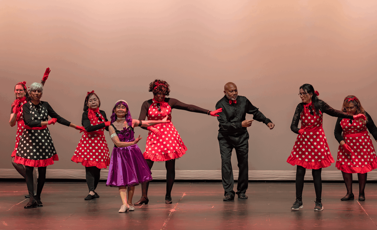 A group of seven dancers performs on a stage, most wearing red polka-dot dresses and red gloves.