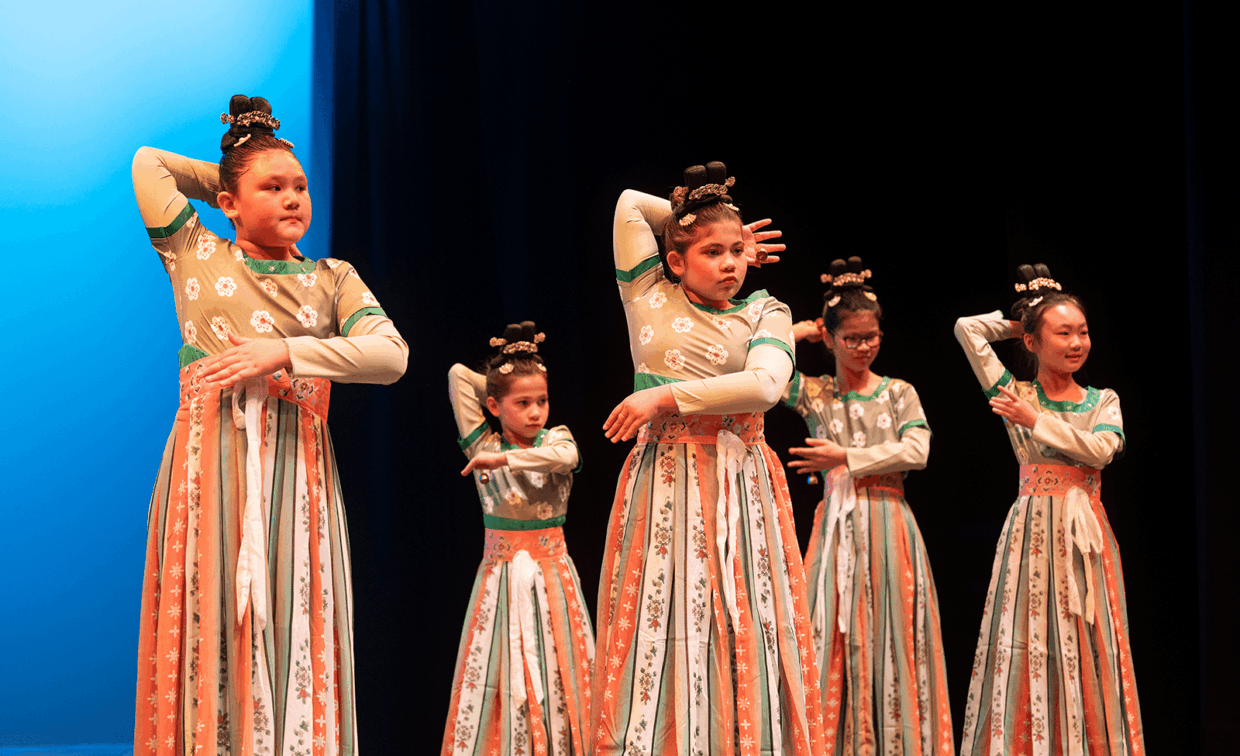 Five dancers in traditional patterned dresses and hair buns perform on a stage with a blue backdrop.