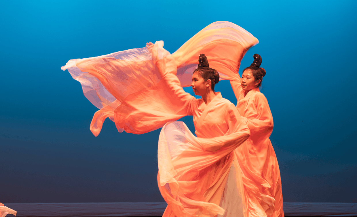 Two dancers in traditional pale orange robes and tall hair buns perform with flowing silk sleeves against a blue background.