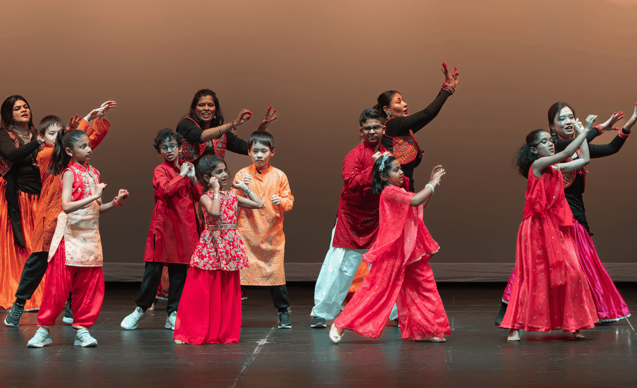 A group of children and adults performing a dance on stage in coordinated red and orange traditional clothing.