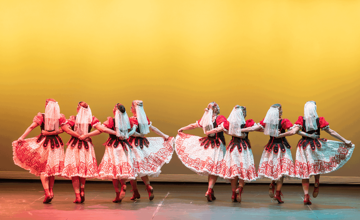 A row of eight dancers in traditional folk costumes and lace head coverings performing on stage against a yellow background.