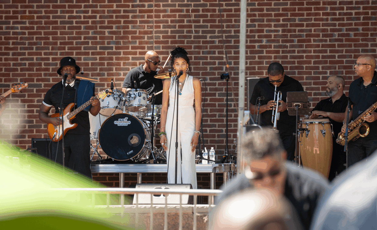 A band performing on an outdoor stage in front of a brick wall, featuring a female lead singer, a bassist, a drummer, a trumpeter, a percussionist, and a saxophonist.