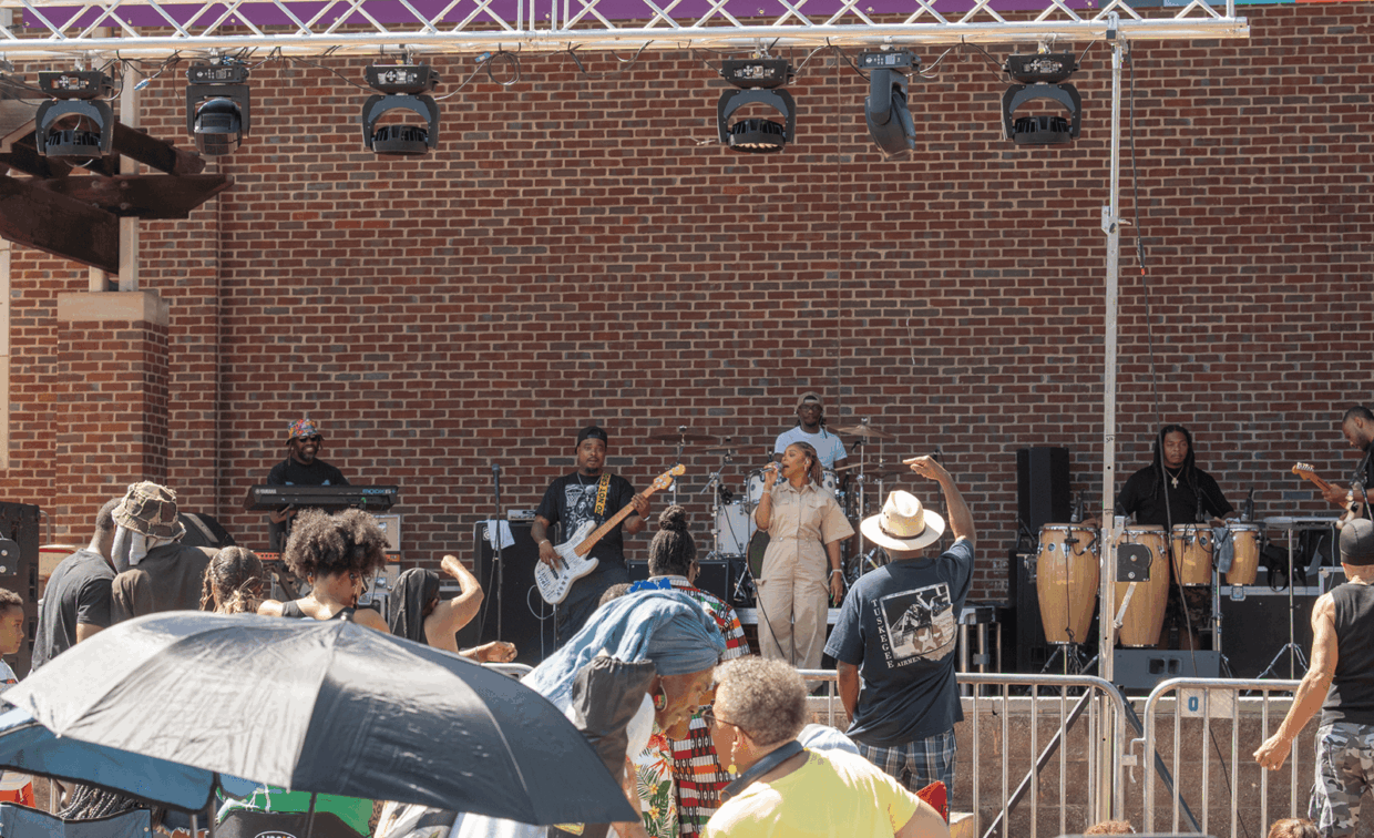 A band performing on an outdoor stage in front of a brick wall, with a crowd watching from behind a metal barricade.