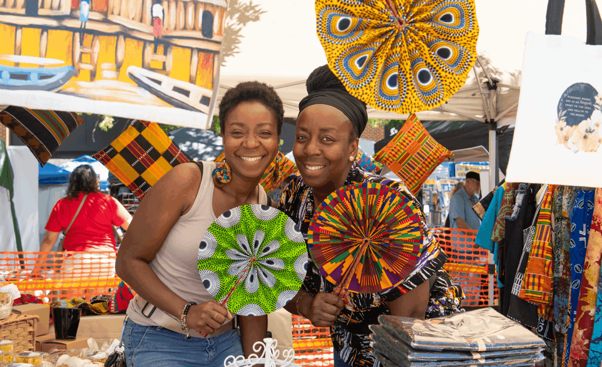Two women smiling behind a market stall display of colorful patterned folding fans and textiles.