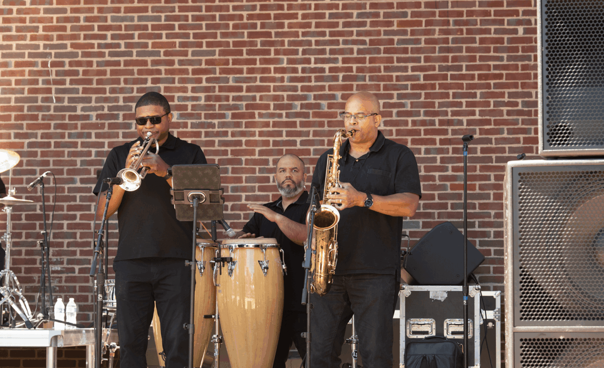 Three musicians in black shirts performing in front of a brick wall: a trumpeter, a conga player, and a saxophonist.