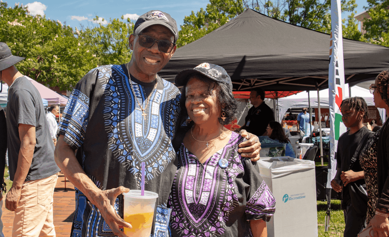 An older man and woman wearing matching patterned shirts and baseball caps pose for a photo at an outdoor community event.
