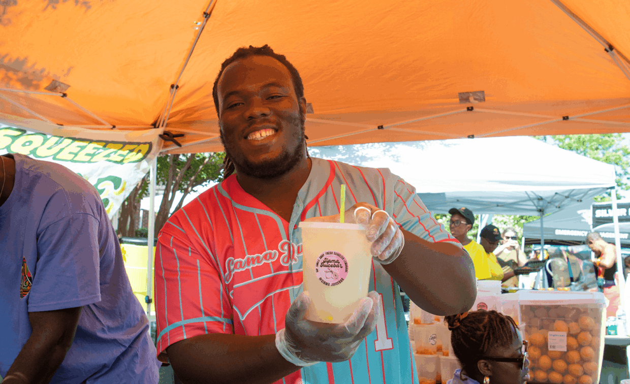 A smiling man in a pink and blue baseball jersey holds out a cup of juice with a “Jama Juicebar” sticker at an outdoor market.