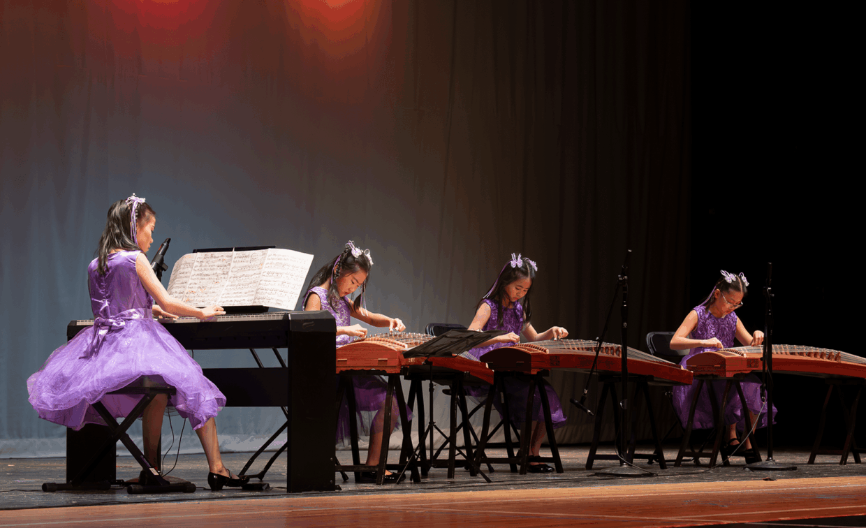 Four girls in matching purple dresses perform on stage, one playing an electronic keyboard and three playing guzheng zithers.
