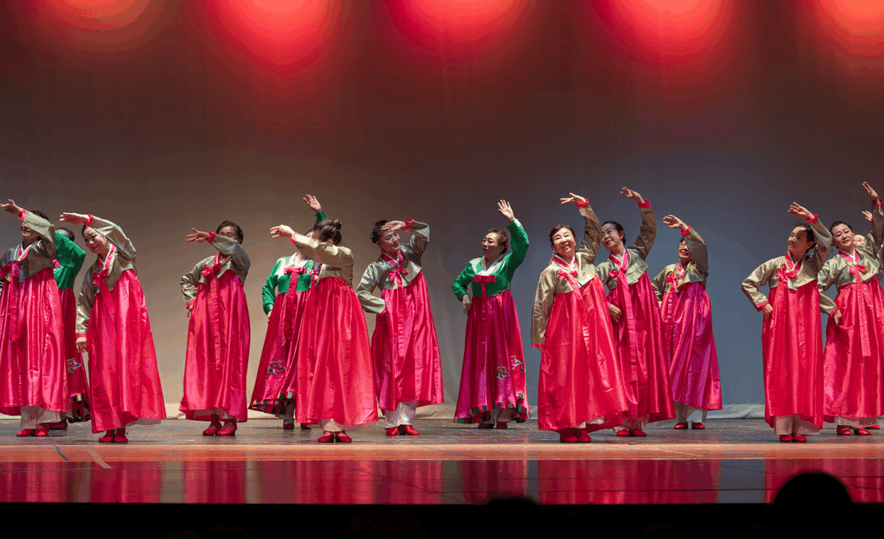 A group of women in traditional Korean hanbok perform a dance on a stage under red spotlights.