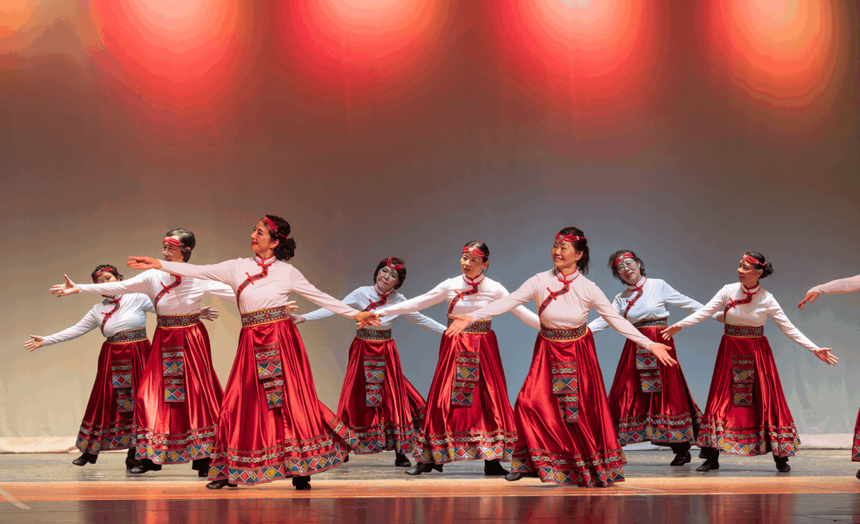 A group of women in white long-sleeved tops and red patterned skirts performing a dance on stage.