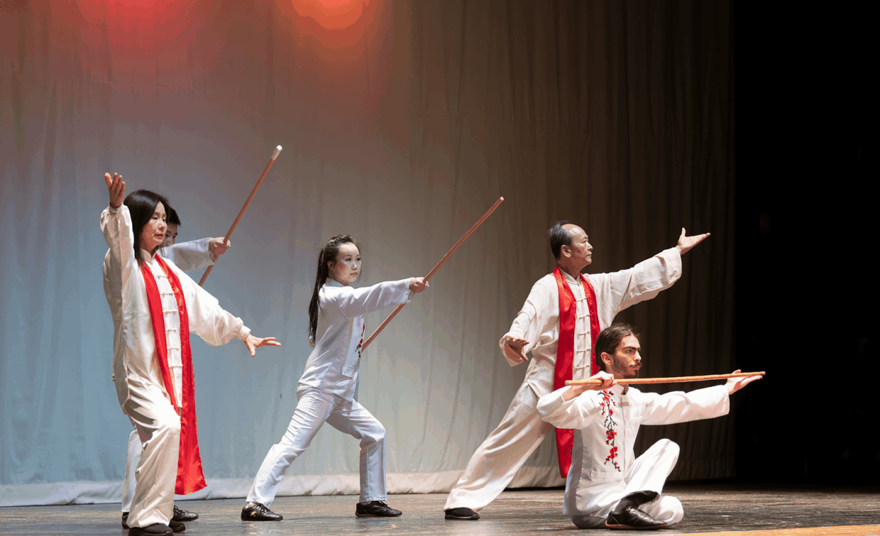 Four performers in white martial arts uniforms with red accents practice tai chi with wooden staffs on a stage.
