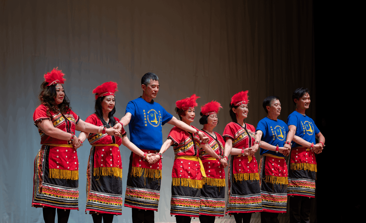 A group of performers wearing traditional red and black costumes and matching blue t-shirts hold hands in a line on stage.