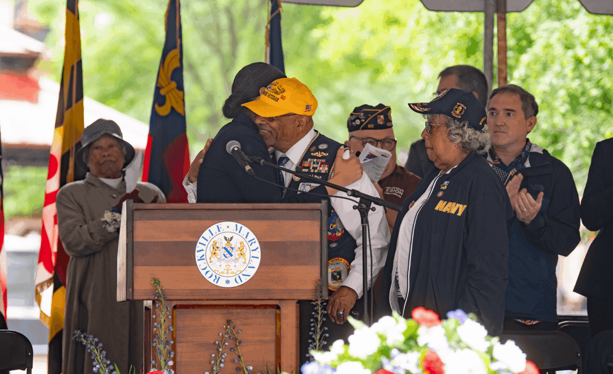 A veteran in a yellow cap embraces a person at a podium featuring the Rockville, Maryland city seal, while a woman wearing a navy jacket and cap looks on.