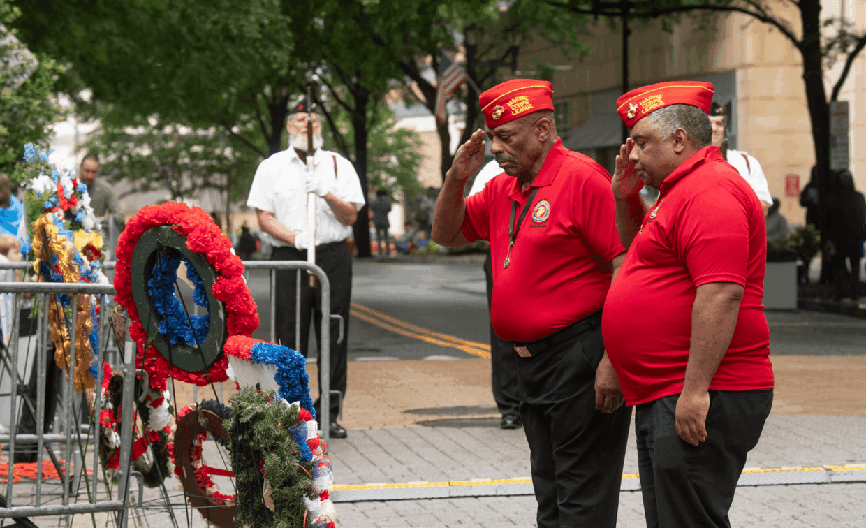 Two Marine Corps League members in red shirts and caps salute at a wreath-laying ceremony.