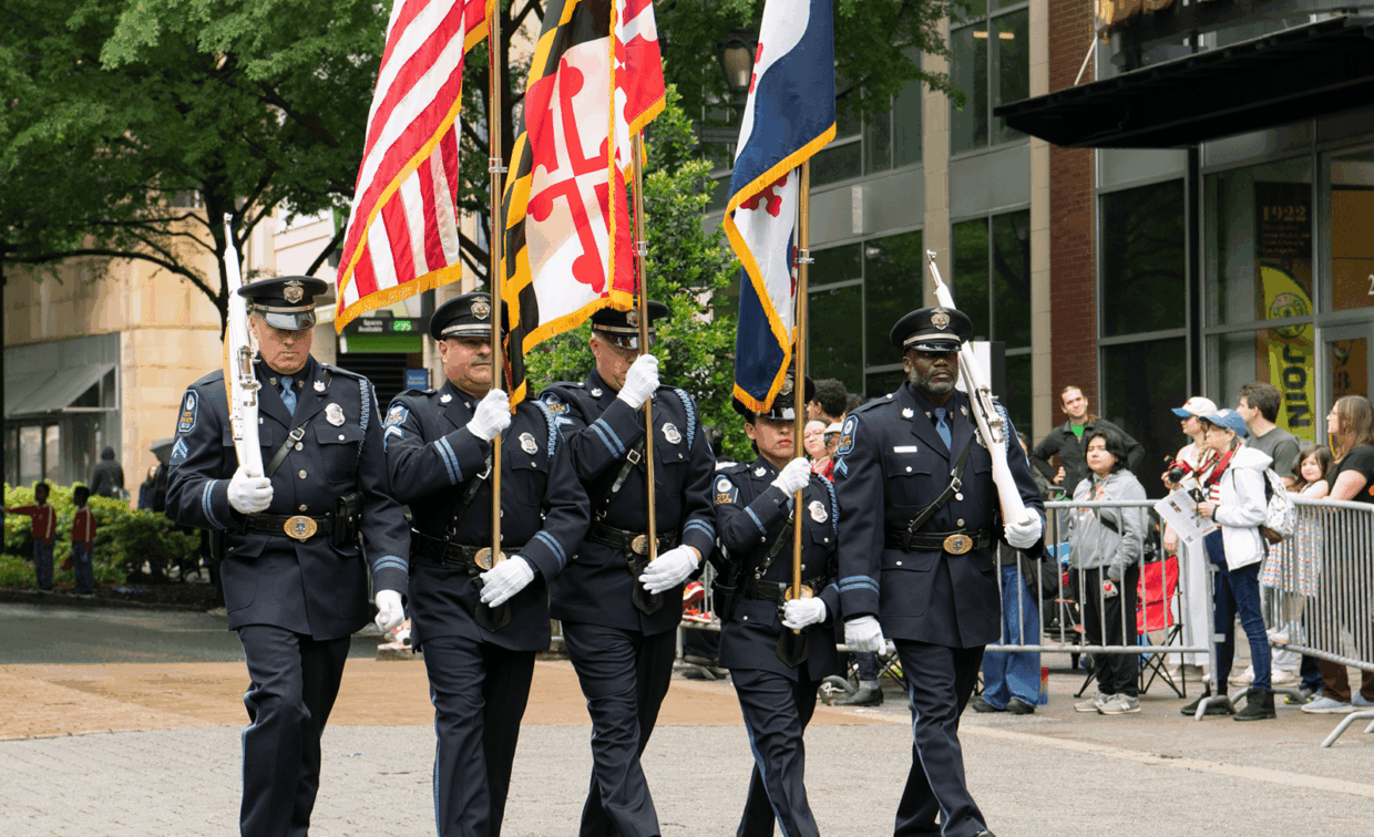 Four police officers in uniform marching in a line and carrying the American, Maryland, and state flags.