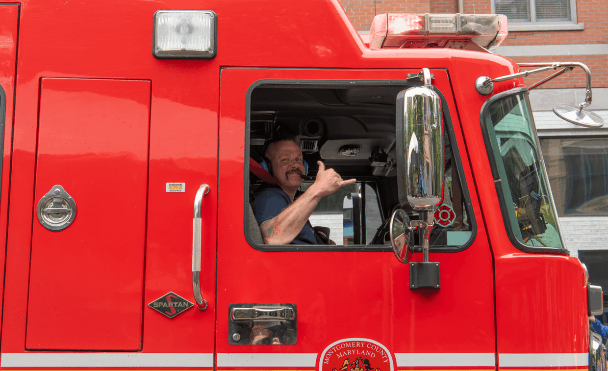 A firefighter in the driver’s seat of a red fire engine makes a shaka hand gesture.