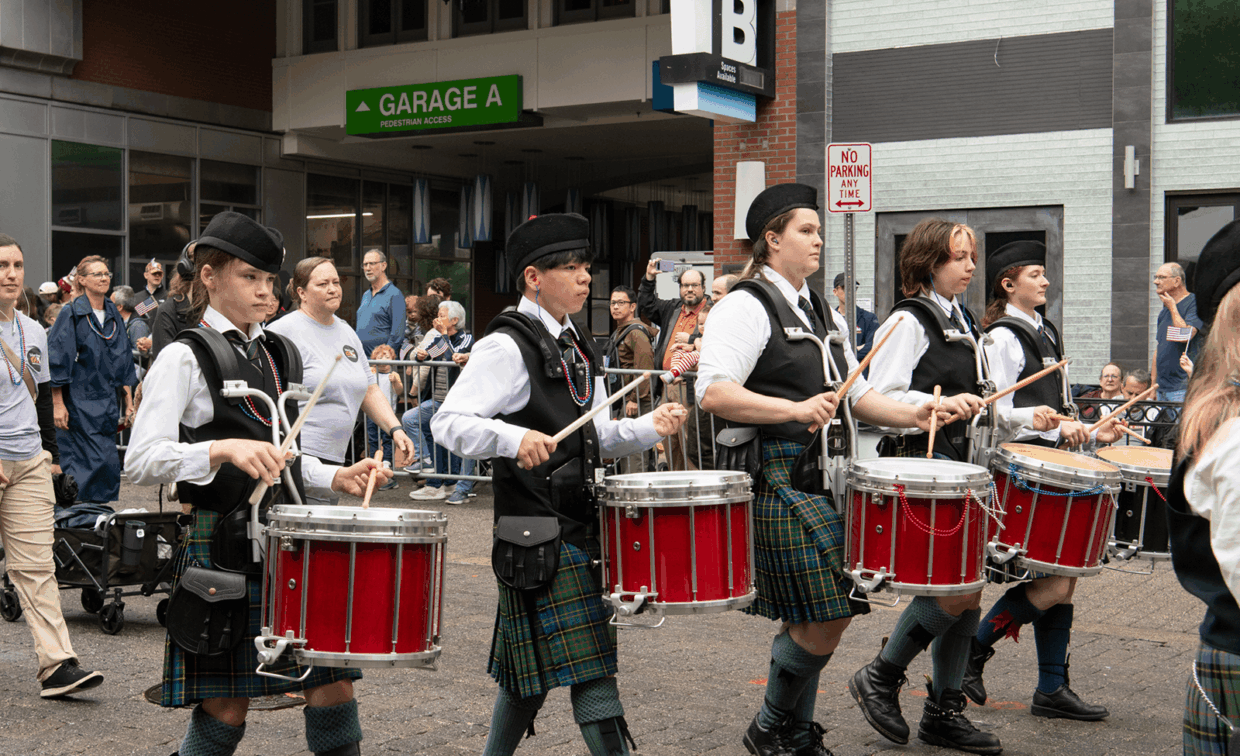A line of four drummers in traditional highland attire—white shirts, black vests, and plaid kilts—marching and playing red drums during a parade.