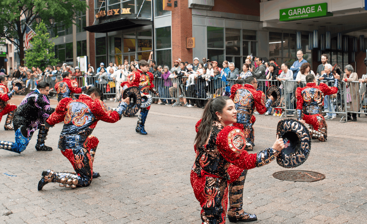 Dancers in matching red and blue sequined costumes perform in a street parade while holding circular hats.