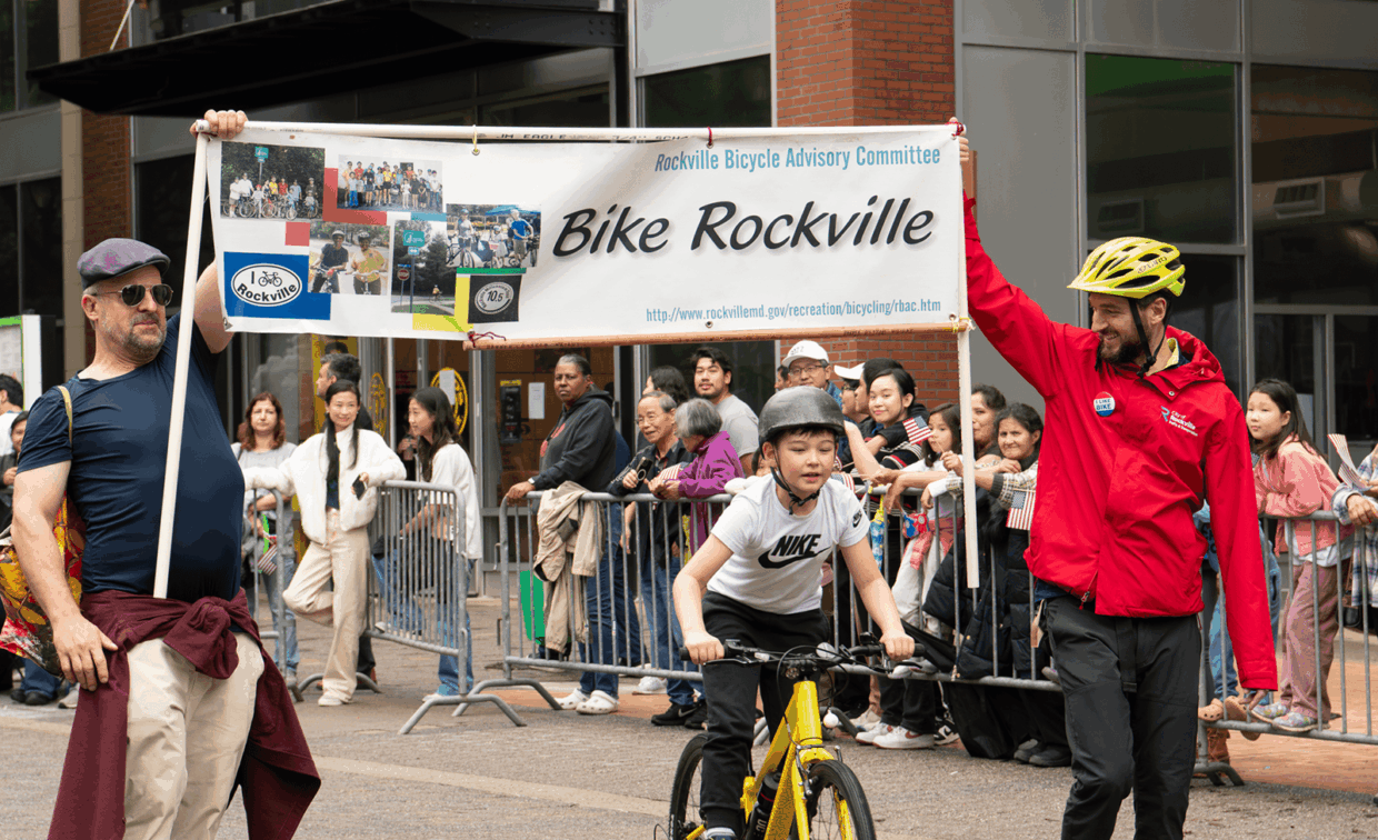 Two men in a parade hold a banner that reads “Bike Rockville,” as a child rides a yellow bicycle between them in front of a crowd.