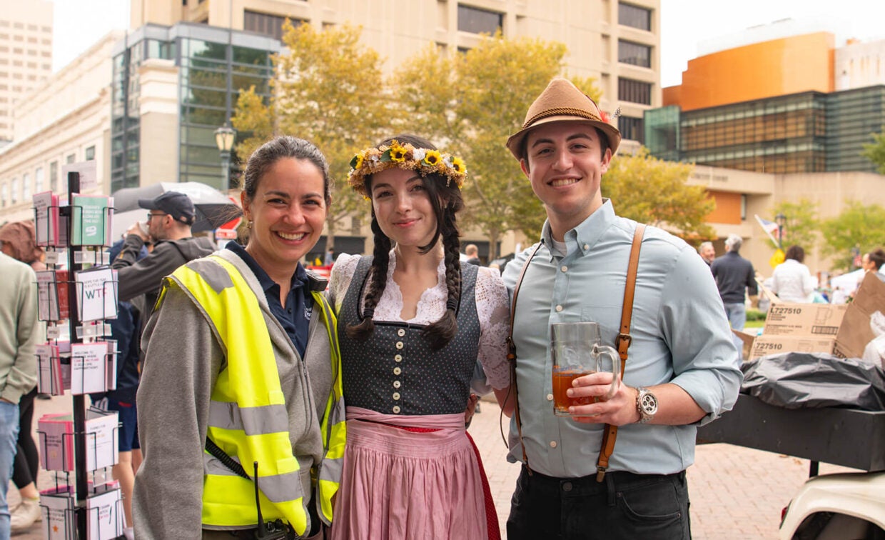 Three people smiling at an outdoor event: a woman in a neon safety vest, a woman wearing a flower crown and traditional Bavarian dress, and a man in a hat and suspenders holding a beer stein.
