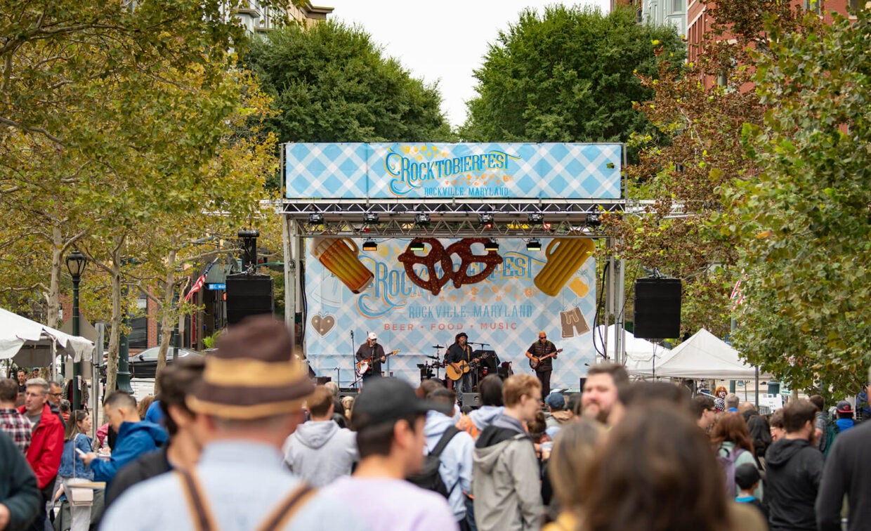 A band performs on a stage decorated with a “Rocktoberfest Rockville, Maryland” banner in front of a crowd.