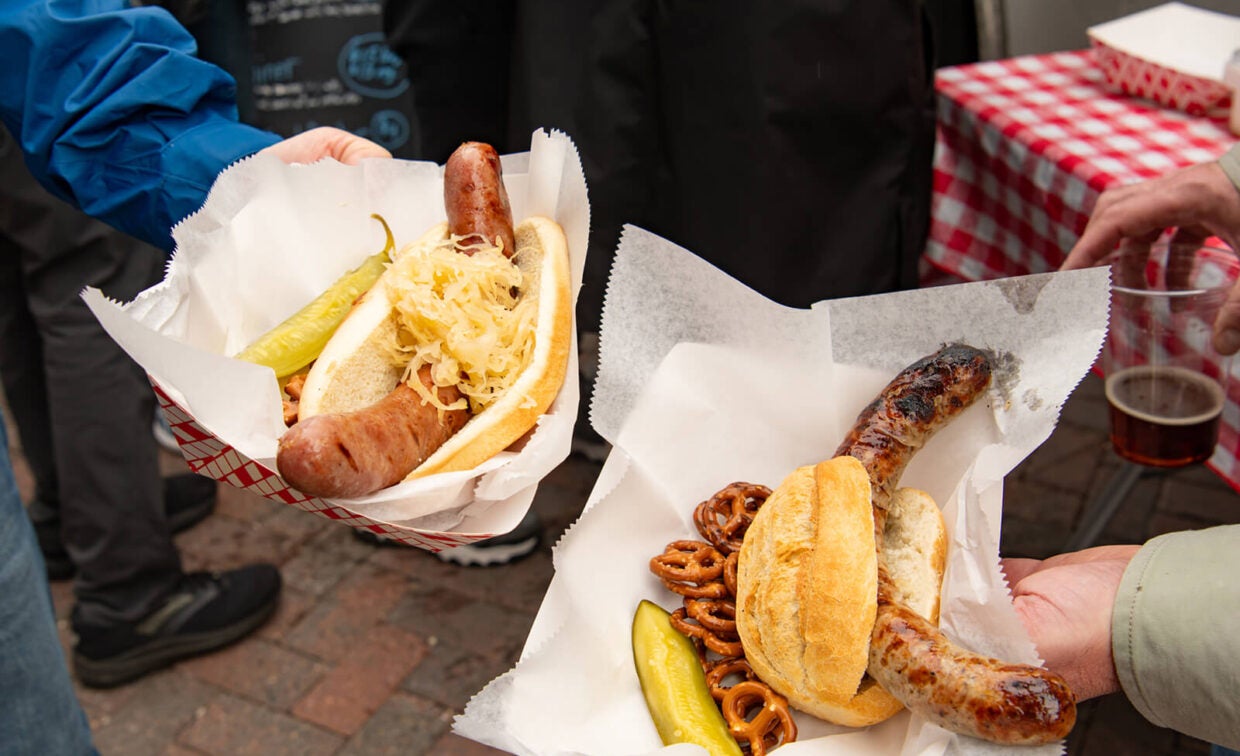 Two people holding paper baskets containing sausages in buns with sauerkraut, a pickle, and pretzels.